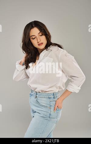 Eine atemberaubende Frau in Übergröße posiert in einem trendigen weißen Hemd und Jeans vor grauem Hintergrund. Stockfoto
