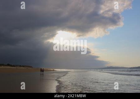 Am späten Nachmittag taucht die September-Sonne durch die schwere dunkle Wolke auf, mit Spaziergängern in der seichten Brandung am Somo Beach Ribamontán al Mar Cantabria Spanien Stockfoto