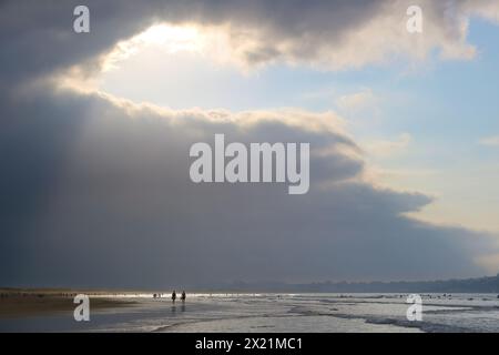 Am späten Nachmittag taucht die September-Sonne durch die schwere dunkle Wolke auf, mit Spaziergängern in der seichten Brandung am Somo Beach Ribamontán al Mar Cantabria Spanien Stockfoto