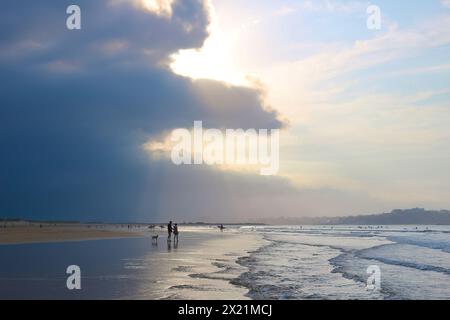 Am späten Nachmittag taucht die September-Sonne durch die schwere dunkle Wolke auf, mit Spaziergängern in der seichten Brandung am Somo Beach Ribamontán al Mar Cantabria Spanien Stockfoto