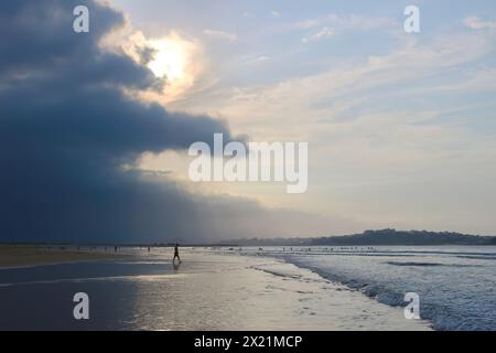 Am späten Nachmittag taucht die September-Sonne durch die schwere dunkle Wolke auf, mit Spaziergängern in der seichten Brandung am Somo Beach Ribamontán al Mar Cantabria Spanien Stockfoto