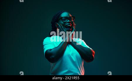 Die überraschte, aufgeregte junge afroamerikanische Frau hält die Hände auf Wangen der Emotionen im Neonlicht vor dem Hintergrund des Gradienten-Studios. Stockfoto