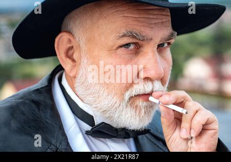 Nahaufnahme des seriösen, stilvollen Hipster-Großvaters im Ledermantel, der draußen raucht. Älterer Großvater, Opa-Rentner, Rentner-Konzept. Stockfoto
