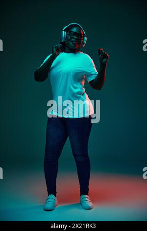 Entspannte afroamerikanische Frau, die Musik in Kopfhörern hört und im roten Neonlicht vor dem Hintergrund des Studiostudios tanzt. Stockfoto