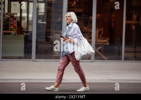 Eine ältere Frau mit kurzen grauen Haaren geht die Stadtstraße entlang, trägt modische Hosen, weißes Hemd, Turnschuhe und Tragetasche, die Stil und Konf ausstrahlt Stockfoto