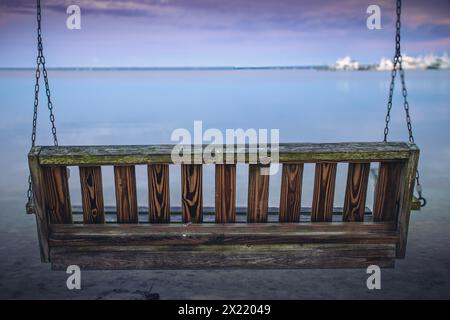 Eine Holzschaukel hängt an Ketten an der Küste des Ozeans. Moos und Witterungseinflüsse deuten darauf hin, dass sie den Elementen ausgesetzt sind. Stockfoto