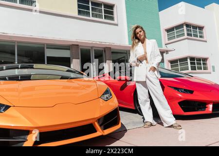 Eine junge blonde Frau, die selbstbewusst neben zwei eleganten Sportwagen in Miami steht. Stockfoto