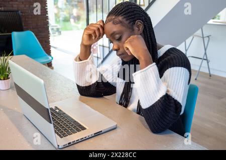 Eine afroamerikanische Frau mit geflochtenen Haaren ist tief in ihre Arbeit vertieft und sitzt an einem modernen Schreibtisch. Ihr Ausdruck spiegelt Konzentration oder Besorgnis wider, während sie auf ihren Laptop-Bildschirm starrt. Sie trägt einen lässigen, schwarz-weiß gestreiften Strickpullover in einem gut beleuchteten Zimmer mit zeitgemäßer Design-Ästhetik. Nachdenkliche Afroamerikanerin, die am Laptop arbeitet. Hochwertige Fotos Stockfoto