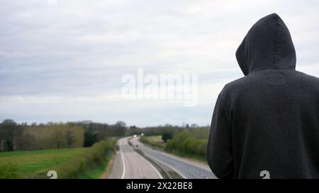 Eine geheimnisvolle Kapuzenfigur, zurück zur Kamera. Blick hinunter auf eine leere Autobahn auf dem Land. Mit Kopierraum Stockfoto