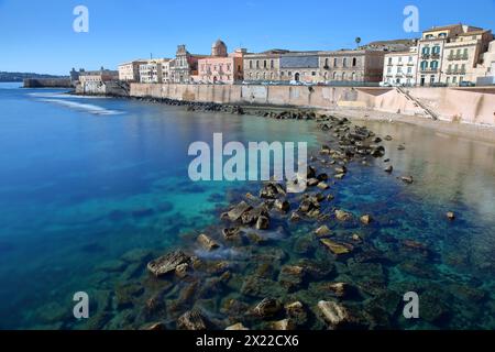 Die östliche felsige Küste der Insel Ortigia, Syrakus, Sizilien, Italien, mit klarem und farbenfrohem Wasser und Castello Maniace (Schloss Maniace) im Hintergrund Stockfoto