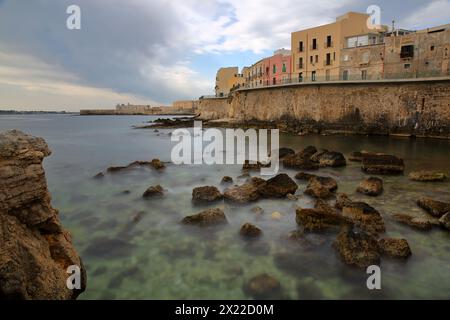 Die östliche felsige Küste der Insel Ortigia, Syrakus, Sizilien, Italien, mit klarem und farbenfrohem Wasser und Castello Maniace (Schloss Maniace) im Hintergrund Stockfoto