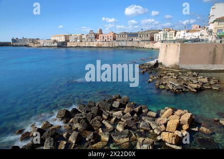 Die östliche felsige Küste der Insel Ortigia, Syrakus, Sizilien, Italien, mit klarem und farbenfrohem Wasser und Castello Maniace (Schloss Maniace) im Hintergrund Stockfoto