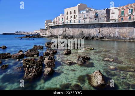 Die östliche felsige Küste der Insel Ortigia, Syrakus, Sizilien, Italien, mit klarem und farbenfrohem Wasser und Castello Maniace (Schloss Maniace) im Hintergrund Stockfoto