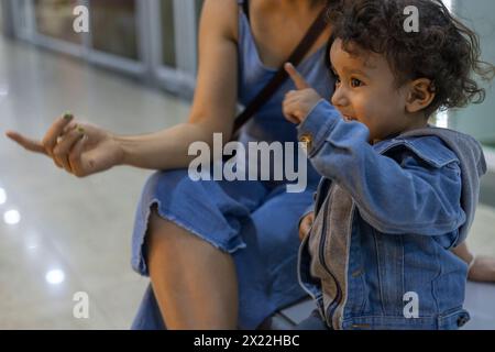 Lateinamerikanischer Junge, der neben seiner Mutter sitzt und mit dem Finger auf etwas zeigt, das er von ihr sehen will. Mutterschaftskonzept Stockfoto