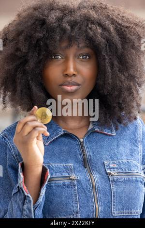 Eine nachdenkliche schwarze Frau mit voluminösem Afro-Haar hält ein Bitcoin-Token hoch und untersucht es genau. Ihre Denim-Jacke ist ein lässiger, aber trendiger Stil, wobei die Münze die Kreuzung von Mode und der wachsenden Welt der Kryptowährung symbolisiert. Neugierige Frau, die Bitcoin im Denim-Outfit hält. Hochwertige Fotos Stockfoto