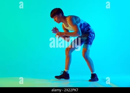 Sportlicher junger Mann in blauer Uniform, in Wrestling-Position, bereit, gegen blauen Hintergrund im Neonlicht zu kämpfen Stockfoto