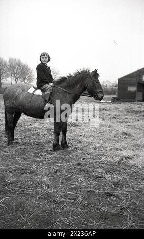 1970er Jahre, historisch, ein junges Schulmädchen, das ihre Schuljacke trägt, auf einem Pferd auf einem Feld sitzt, in einem halbländlichen Reitzentrum, während sie reiten lernt, England, Großbritannien. Stockfoto