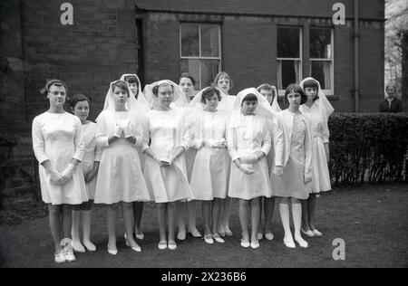 1970er Jahre, historische, katholische Schulmädchen in weißen Kleidern und Haarschleiern, die nach ihrer ersten heiligen Kommunion für ein Foto stehen, England, Großbritannien. Stockfoto