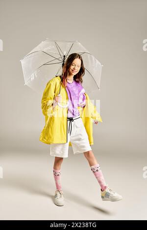 Ein stilvolles Teenager-Mädchen in einem gelben Regenmantel hält fröhlich einen Regenschirm und strahlt lebendige Energie und Positivität aus. Stockfoto