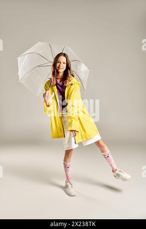 Ein wunderschönes Teenager-Mädchen in einem stilvollen gelben Regenmantel posiert fröhlich und hält an einem regnerischen Tag einen bunten Regenschirm. Stockfoto