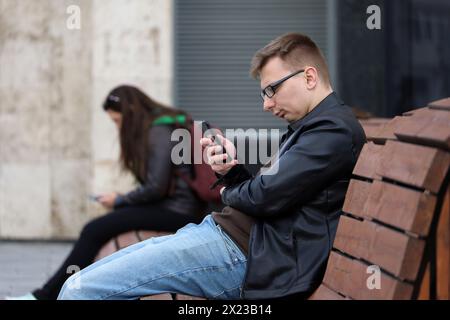 Leute, die Smartphones benutzen, während sie auf einer Holzbank im Frühlingspark sitzen, Mann mit Brille im Vordergrund Stockfoto