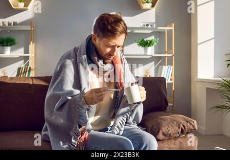 Kranker junger Mann, der auf dem Sofa sitzt, mit Thermometer in Karo gewickelt Stockfoto