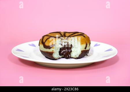 Mit Schokolade gefüllter Donut mit herausgenommenem Biss, und sprudelnder Schokoladenfüllung, auf rosa Hintergrund und hübschem Teller Stockfoto