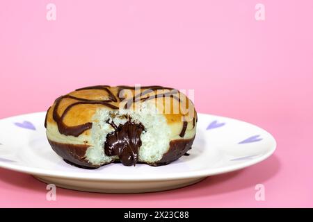 Mit Schokolade gefüllter Donut mit herausgenommenem Biss, und sprudelnder Schokoladenfüllung, auf rosa Hintergrund und hübschem Teller Stockfoto