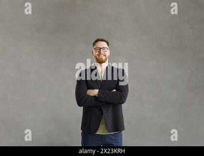 Porträt eines glücklichen jungen Mannes in Brille, die Arme gefaltet vor grauem Atelierhintergrund Stockfoto