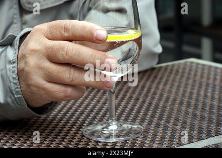 Eine männliche Hand hält ein Weinglas mit Wasser und eine Scheibe Zitrone im Café draußen. Stockfoto