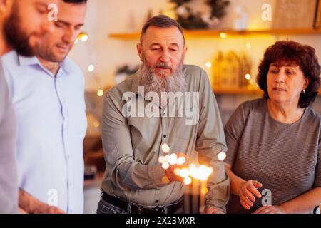 Ein reifes Ehepaar und ihre erwachsenen Söhne feiern zu Hause Geburtstag. Ein älterer Mann mit Bart bläst Kerzen aus. Ein festlicher Kuchen mit Feuer Stockfoto