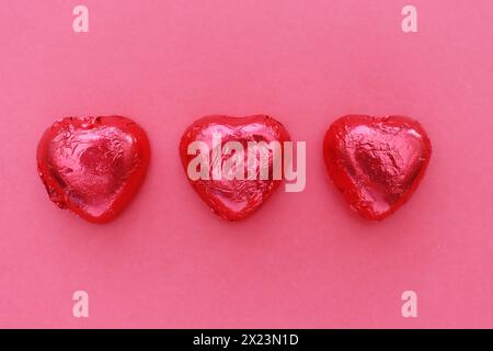 Drei herzförmige Pralinen in roter Folie auf rotem Hintergrund, Draufsicht. Stockfoto