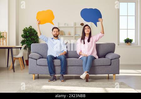 Das junge Paar sitzt auf dem Sofa und jedes von ihnen hält eine farbige Sprechblase. Stockfoto