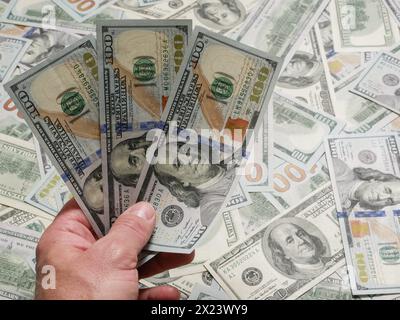 Menschliche Hand hält Banknoten 100 US-Dollar vor dem Hintergrund vieler lügender Dollarscheine. US-Banknoten halten, Nahaufnahme der Hände. Stockfoto