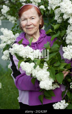 Außenporträt einer alten Seniorin. Schöne ältere Frau lächelt vor dem Hintergrund blühender weißer Flieder im Frühlingspark. Stockfoto