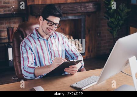 Foto von nettem jungen Geschäftsmann, der Clipboard Docs unterzeichnet, gestreiftes Hemd Loft Innenraum Büro drinnen trägt Stockfoto