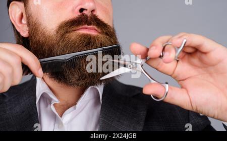 Friseur-Verfahren. Professionelle Bartpflege. Stilvoller bärtiger Mann im Anzug schneidet seinen Bart und Schnurrbart mit Schere und Kamm. Salon für Männer Stockfoto