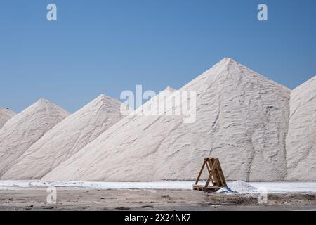 Imposante weiße Salzhaufen bilden einen markanten Kontrast unter einem klaren blauen Himmel, mit einer Holzstruktur im Vordergrund für Skala. Hochwertige Fotos Stockfoto