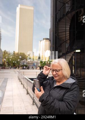 Eine leitende blonde Geschäftsfrau schaut auf das Handy mit einer Brille, um tagsüber im Winter auf der Straße ihre Sehkraft zu überprüfen Stockfoto