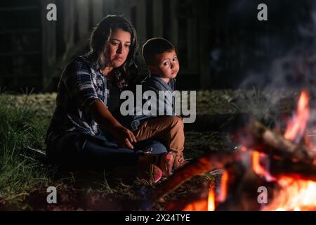 Junge lateinfrau, die ihren Sohn trägt, sitzt nachts vor einem Lagerfeuer und sieht traurig und gelangweilt aus. Stockfoto