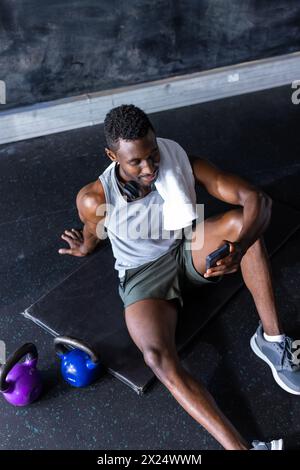 Afroamerikanischer junger männlicher Athlet, der im Fitnessstudio sitzt, Smartphone hält, mit Kettlebells trainiert Stockfoto