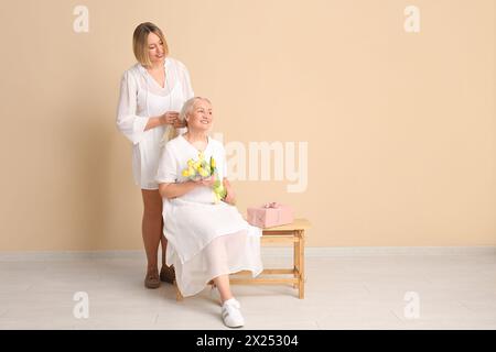 Reife Frau mit ihrer Tochter, Tulpenstrauß und Geschenkbox, die auf einer Bank in der Nähe einer beigefarbenen Wand sitzt. Internationaler Frauentag Stockfoto