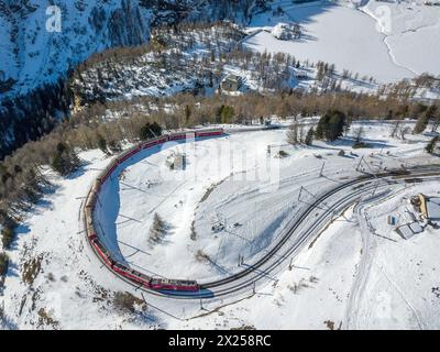 Luftbild eines roten Zuges, der auf dem Gleis der Rhätischen Bahn vorbeifährt, mit der berühmten engen 180°-Kurve auf der hohen Alp Grum über dem Berina-Pass Stockfoto