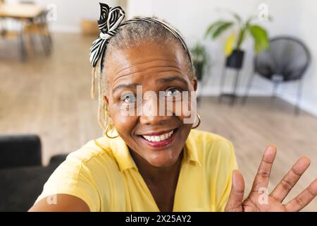 Schwarze Seniorin in gelbem Hemd gibt bei Videoanrufen zu Hause ein Friedenszeichen Stockfoto