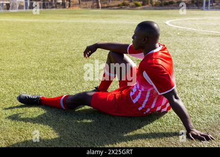 Afroamerikanischer junger männlicher Athlet, der draußen auf dem Rasen sitzt und rote Fußballuniform trägt Stockfoto