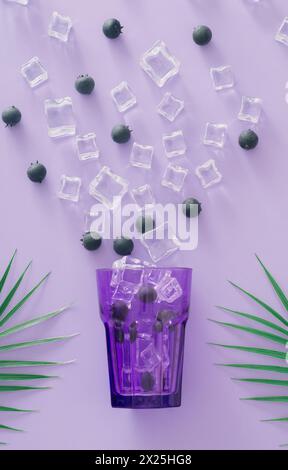 Kreatives exotisches Layout mit lila Glas, frischen Blaubeeren, Eiswürfeln und tropischen grünen Palmblättern auf lila Hintergrund. Minimales Sommergetränk Stockfoto