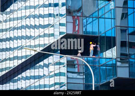 Zwei Männer mit Schutzhüten (Glaser) arbeiten an einem großen Stück Glas, Teil einer Glasvorhangfassade, auf einem Hochhaus in Singapur Stockfoto