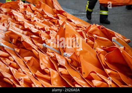 Schweinfurt, Deutschland. April 2024. Ein Ölausleger ist einsatzbereit, während ein Feuerwehrmann vorbeigeht. Die Feuerwehren der Stadt und des Landkreises Schweinfurt und die Wasserrettungseinheiten am Main üben im Katastrophenfall. Es wird davon ausgegangen, dass Öl auf fließendes Wasser austritt. Quelle: Pia Bayer/dpa/Alamy Live News Stockfoto