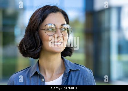 Eine fröhliche Studentin genießt einen sonnigen Tag auf dem Campus mit Brille und lässigem Jeansshirt. Ihr Lächeln spiegelt Selbstvertrauen und Zufriedenheit in einer lehrreichen Umgebung wider. Stockfoto