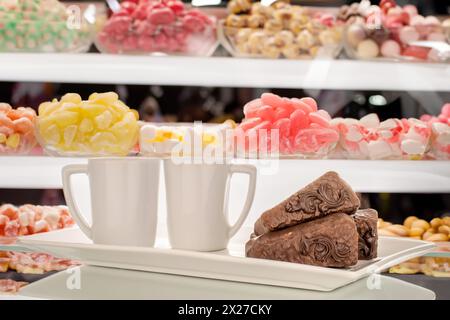 Drei Stücke Schokoladenwaffelkuchen mit zwei Tassen auf einer weißen Keramikplatte, auf dem Hintergrund einer Vitrine mit Süßigkeiten. Stockfoto